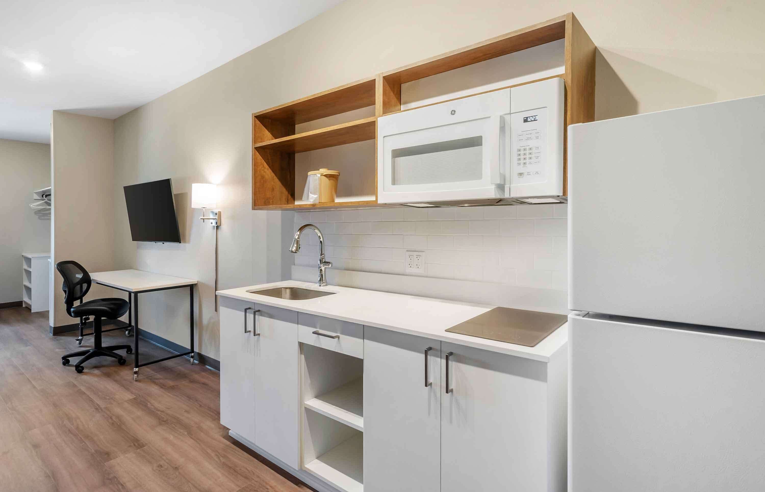 A modern kitchen with white cabinets, a microwave, and a refrigerator. There is a small wooden shelf above the microwave and a desk with a chair to the left.