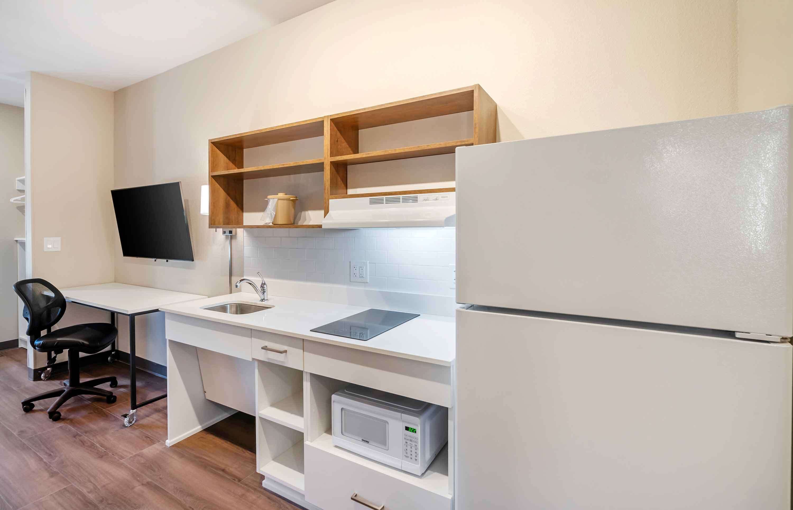 A compact kitchen with a white countertop, a built-in microwave, a refrigerator, and a wooden shelf above the sink.