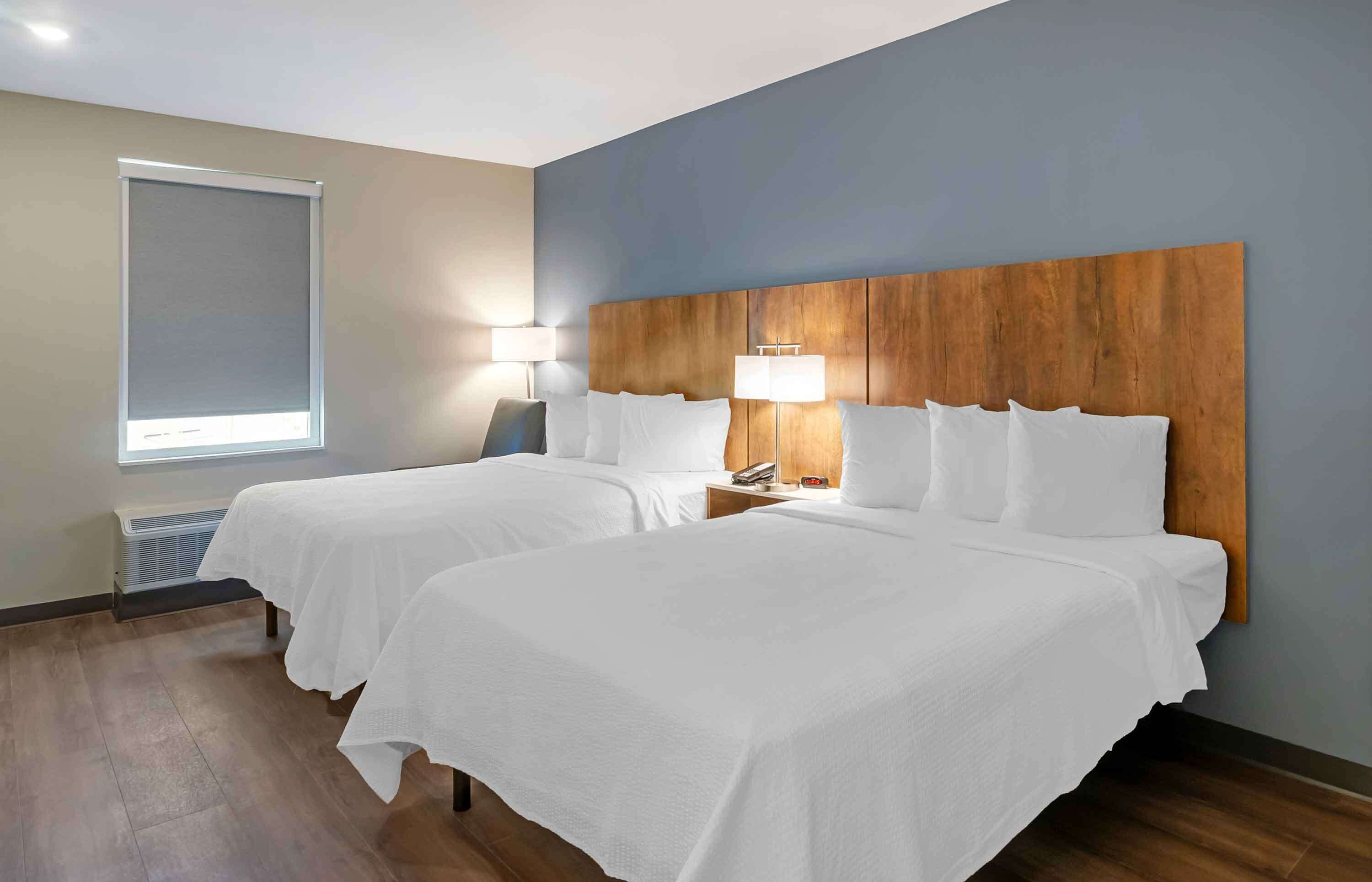 Une chambre d’hôtel avec deux lits, une tête de lit en bois, une fenêtre avec un store et une lampe de chevet.