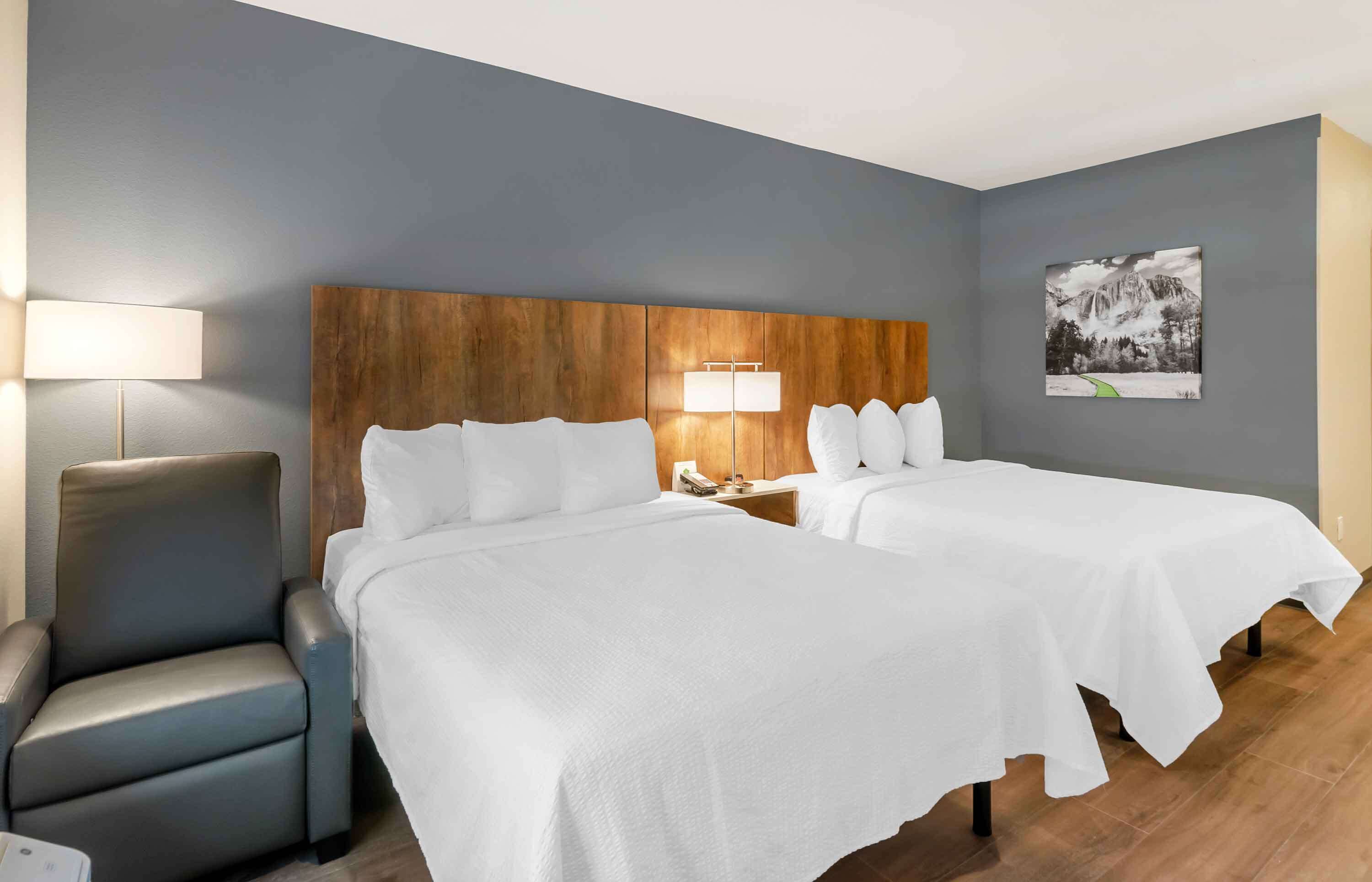 Camera d'albergo con due letti, una testiera in legno, una lampada, una sedia e un quadro incorniciato appeso al muro.