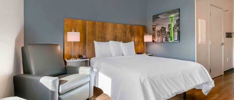 Una habitación de hotel con cama, una silla, una mesita de noche, una lámpara y un cuadro de un paisaje urbano en la pared.