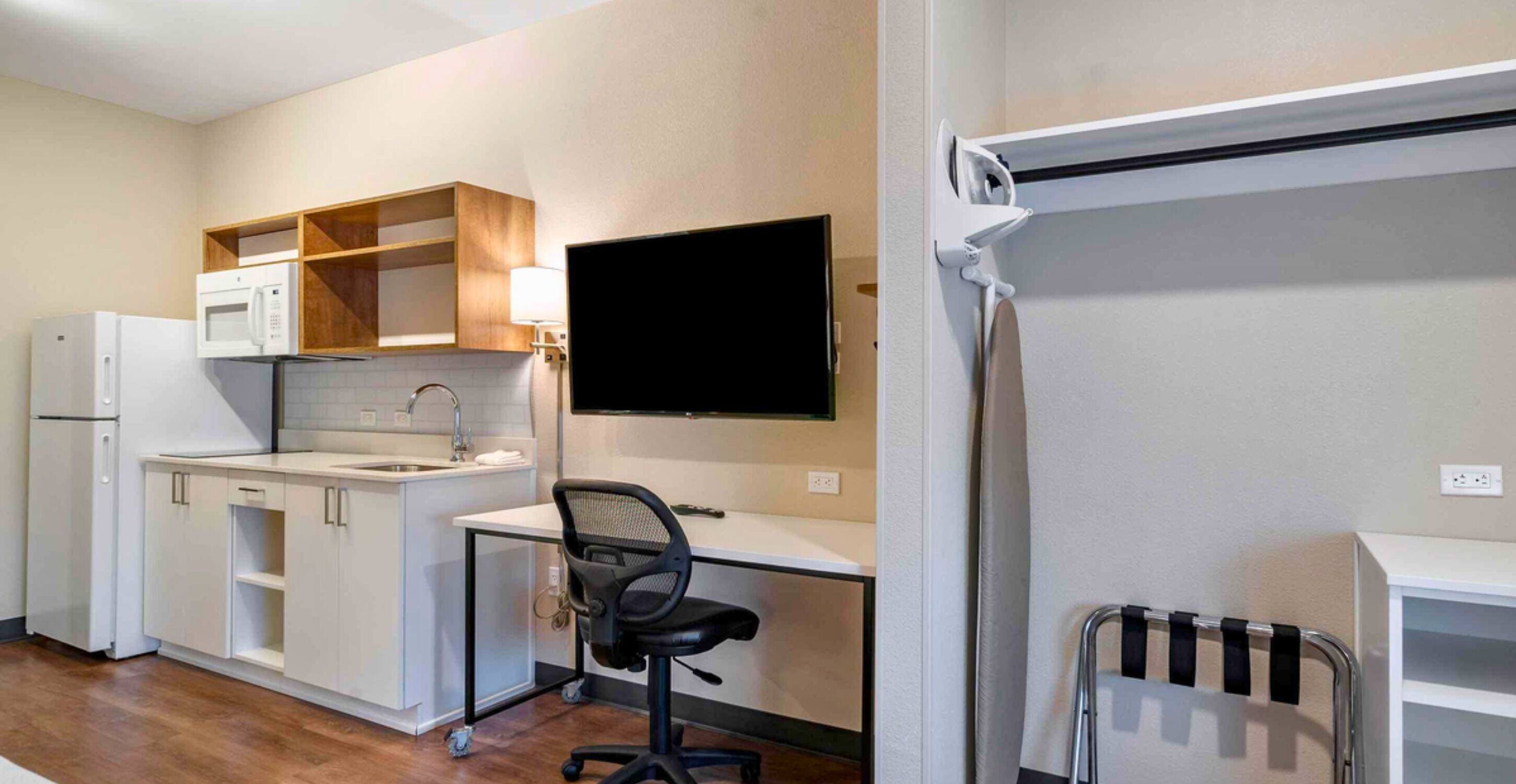 A compact kitchen with a microwave, sink, and cabinets, a small office area with a chair, and a built-in TV.