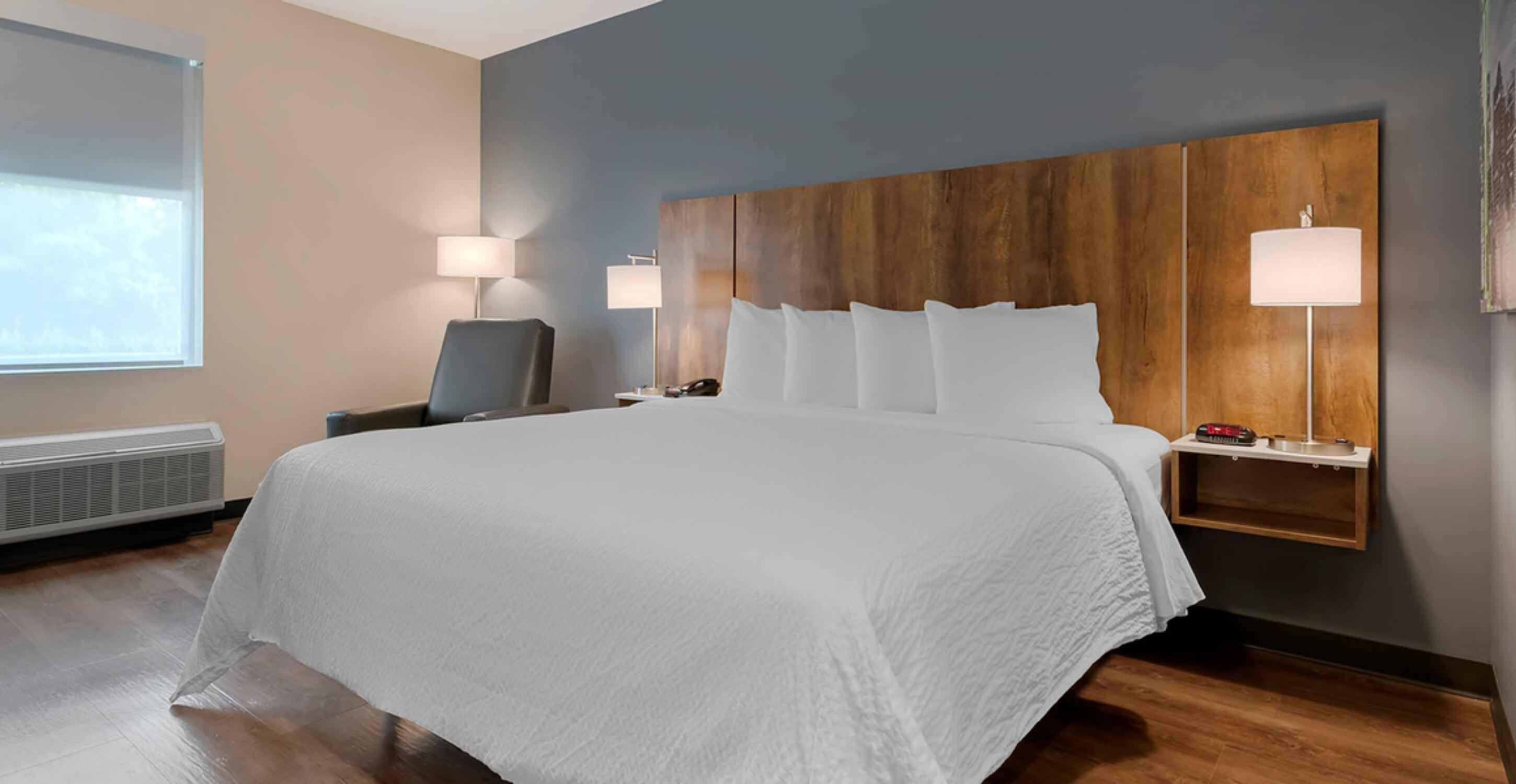 A hotel room with a large bed, a desk, two bedside lamps, and a wall-mounted shelf with a phone and a clock.