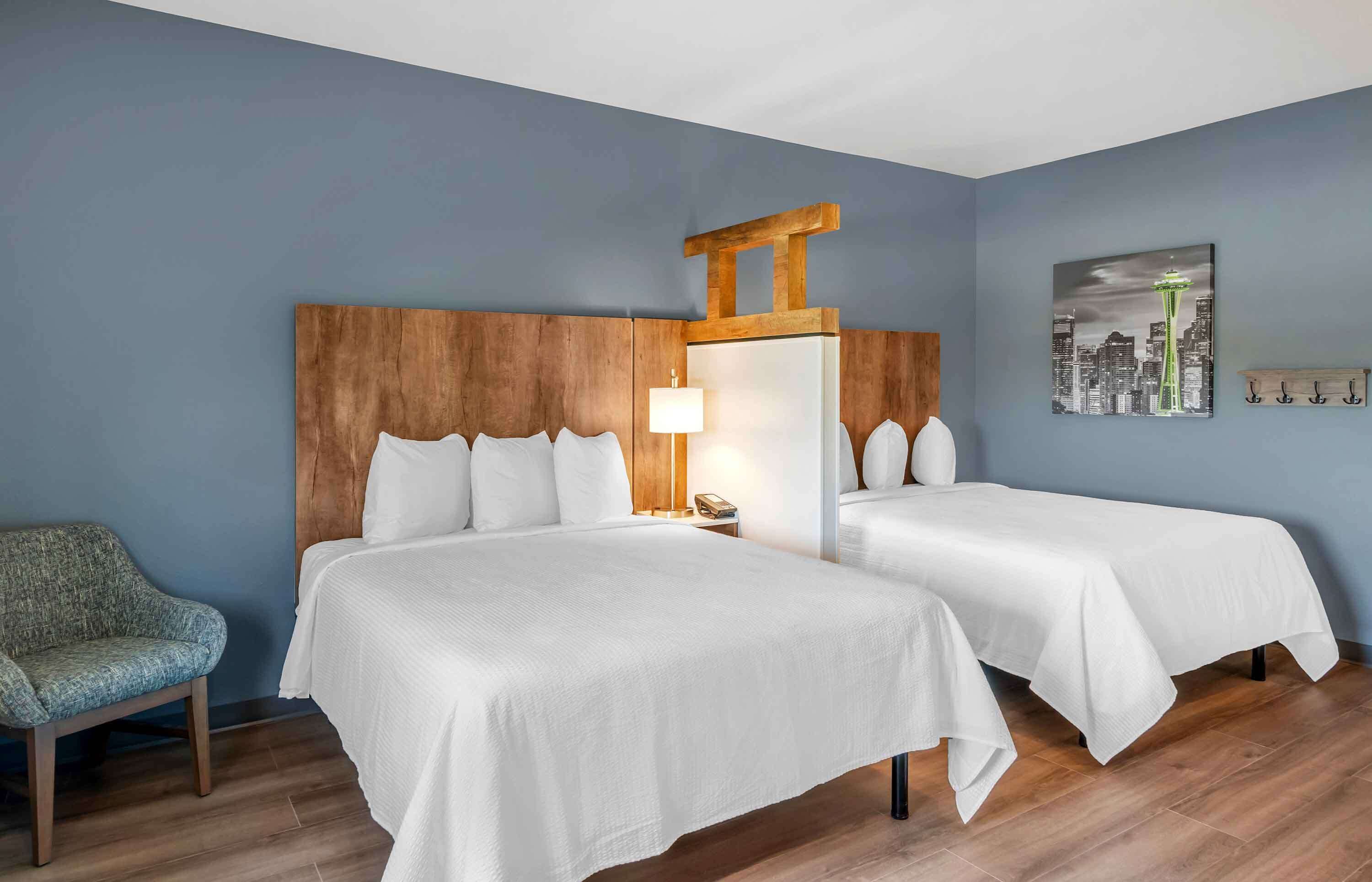 Habitación de hotel con dos camas, cabecera de madera, una silla, una lámpara y un cuadro con una vista urbana.