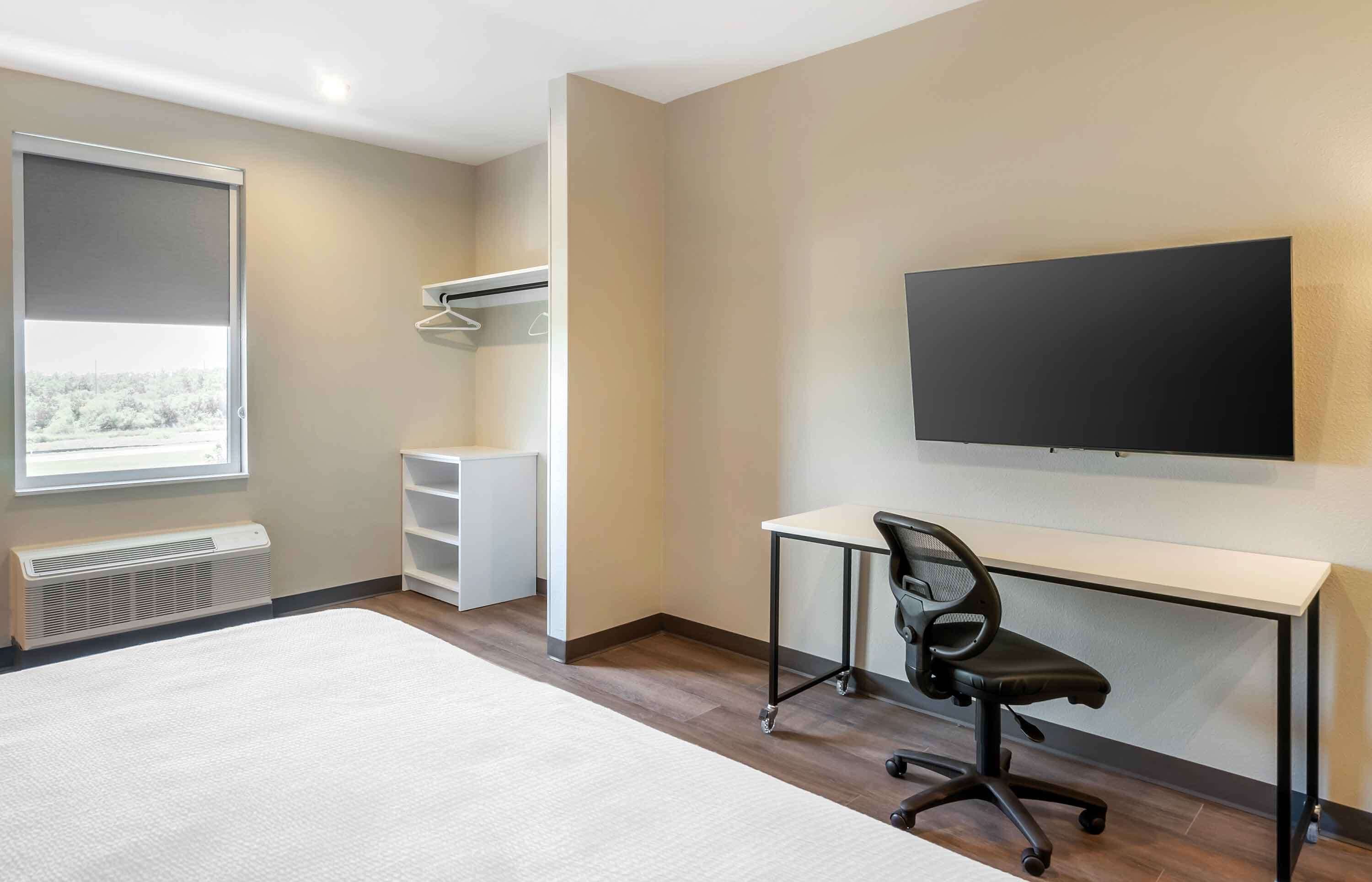 A hotel room with a large bed, a desk with a chair, a TV mounted on the wall, a wardrobe, and a window with a view of trees.