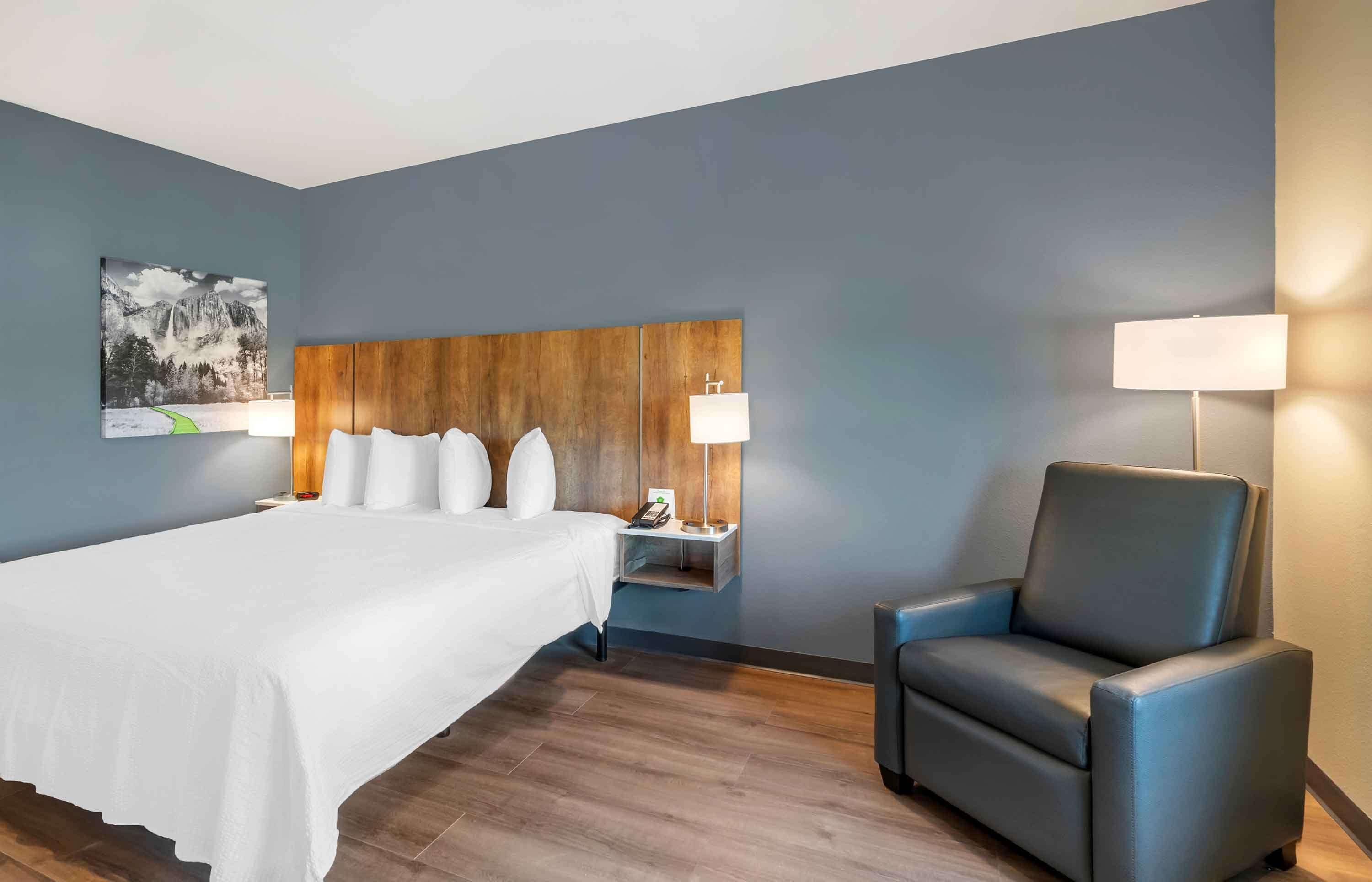 A hotel room with a large bed, a wooden headboard, two bedside tables with lamps, a blue armchair, and a wall-mounted painting.