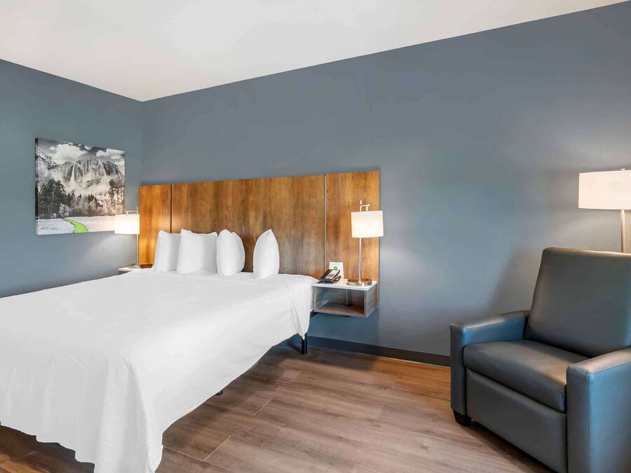 A hotel room with a large bed, a wooden headboard, two bedside tables with lamps, a blue armchair, and a wall-mounted painting.
