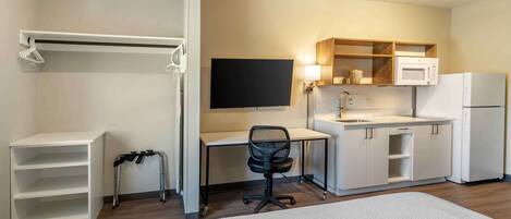 A hotel room with a bed, a desk with a chair, a television, a microwave, a refrigerator, a small kitchen area, a shelf unit, and a walking aid.