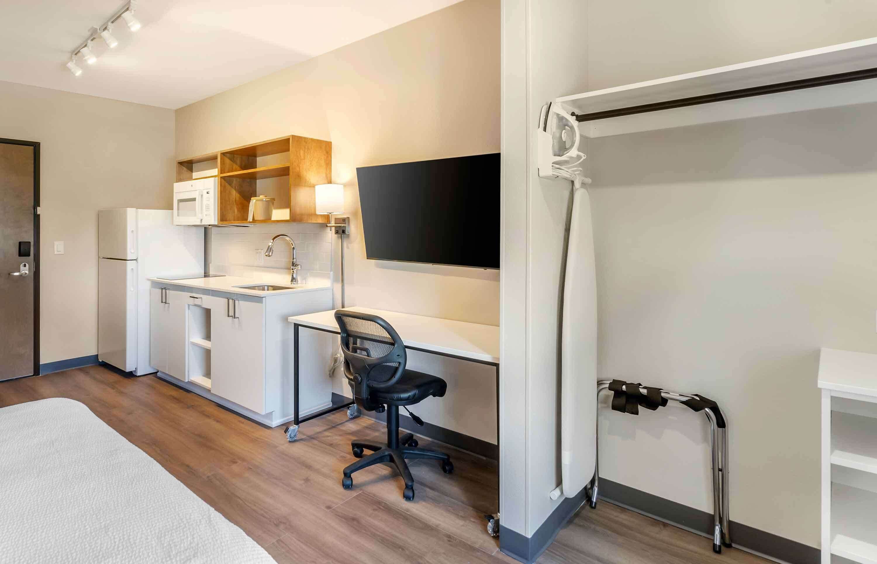 A modern hotel room with a kitchenette, a desk with a chair, a television, and a walk-in closet.