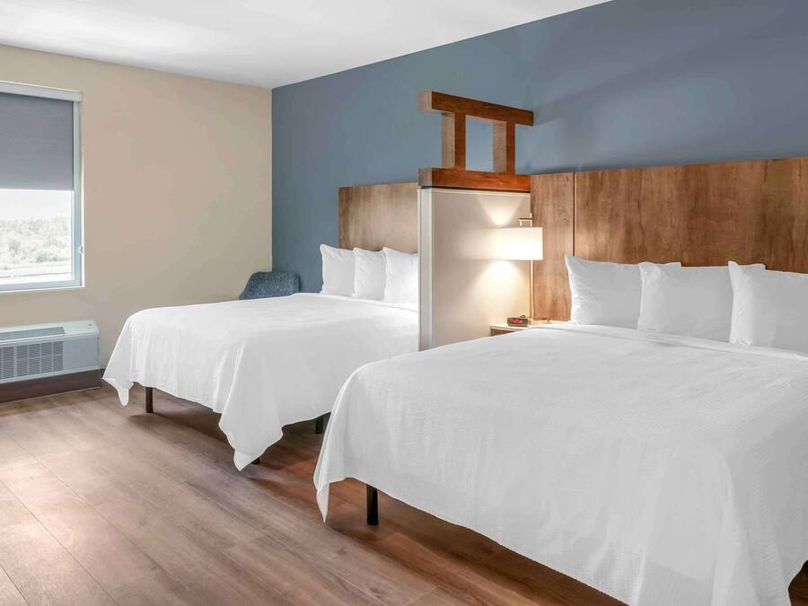 A hotel room with two beds, a wooden headboard, a window with a view, and a wall-mounted air conditioner.