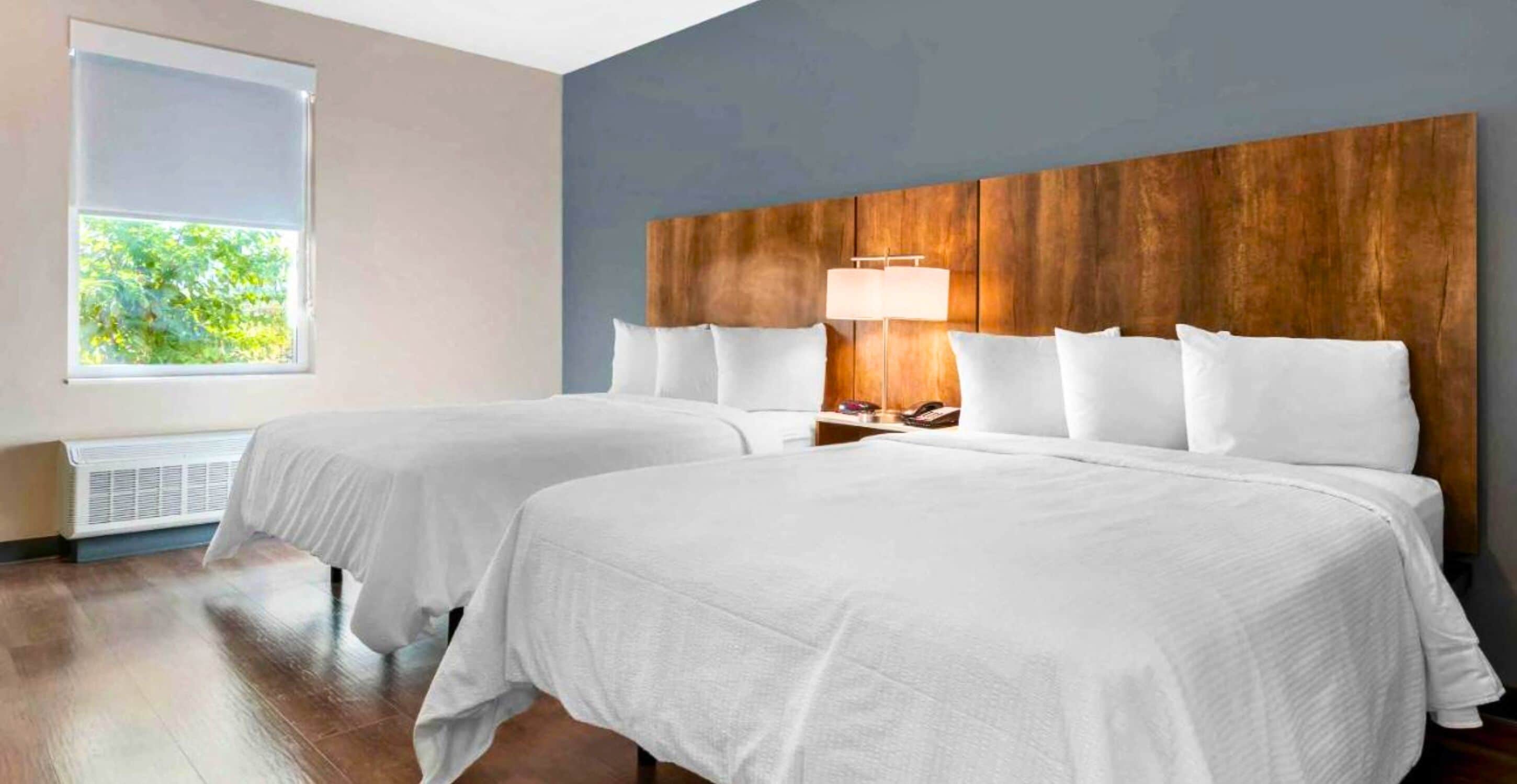 Une chambre d’hôtel avec deux lits, une tête de lit en bois, une lampe de chevet et une fenêtre donnant sur un paysage verdoyant.