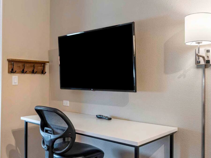 A hotel room with a desk, chair, TV, and a wall-mounted lamp.