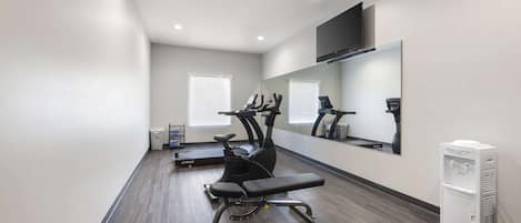Un gimnasio con equipo de ejercicio, una banca, un televisor en la pared y un dispensador de agua.