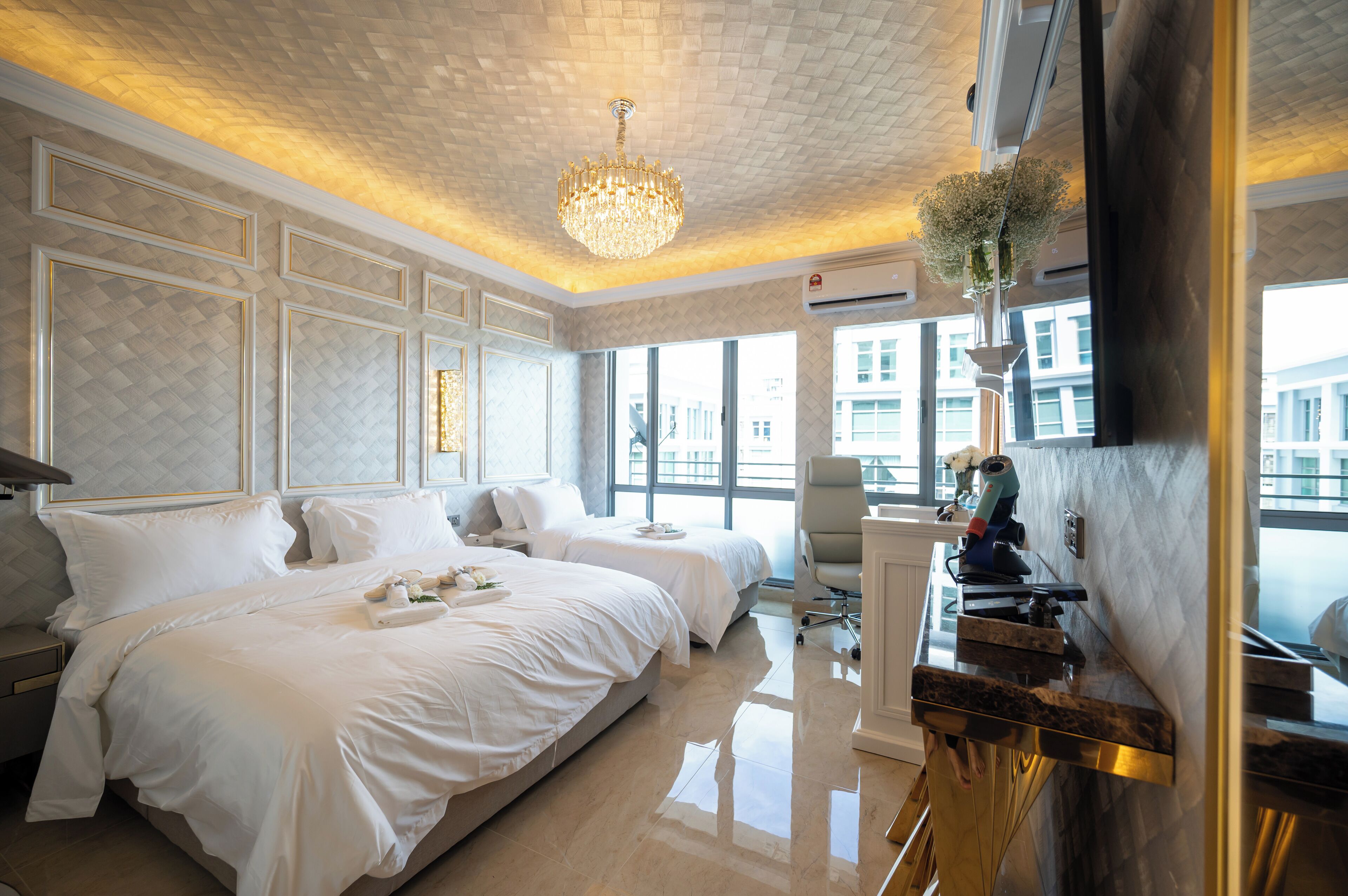 A hotel room with two beds, a large window, a chandelier, and a modern kitchen area.