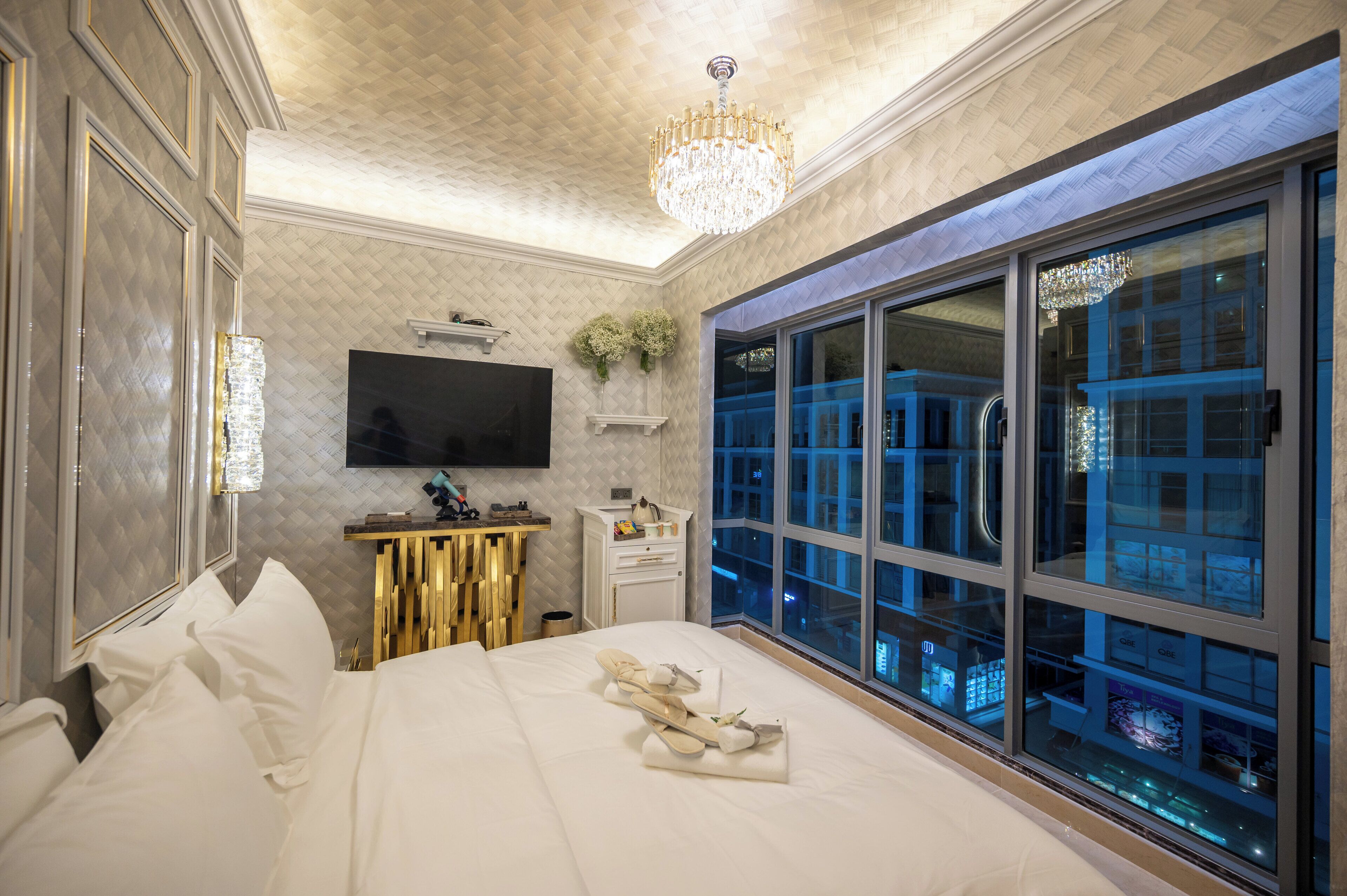 A modern hotel room with a large bed, a TV, a desk with a computer, and a large window with a city view.