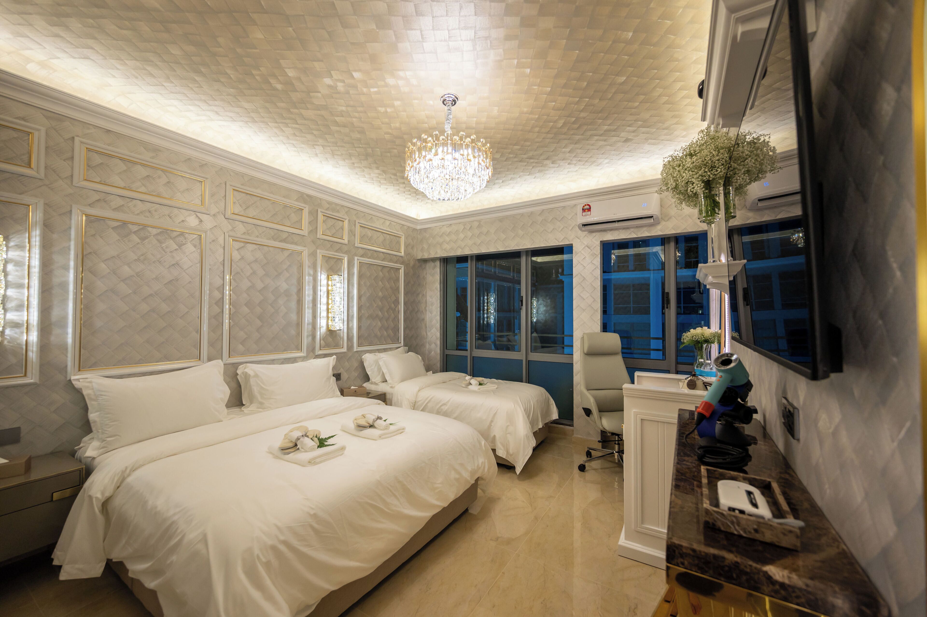 A hotel room with two beds, a desk, a chair, a chandelier, and a large window.