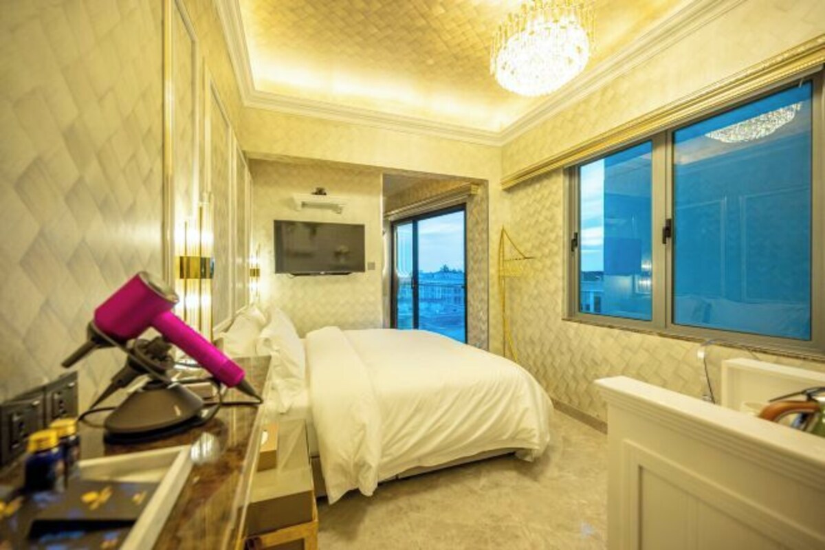 A hotel room with a bed, a desk with a pink hairdryer, a window with a view, and a chandelier.