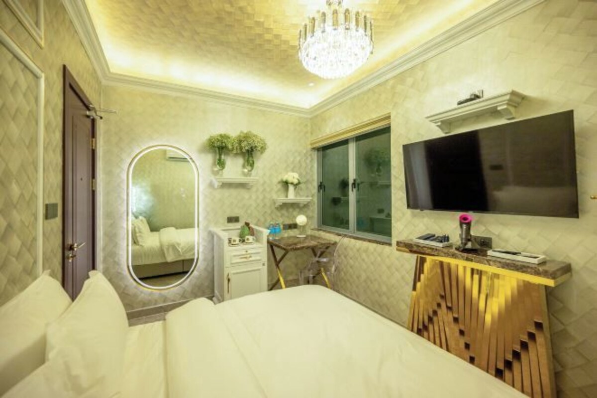 A bedroom with a bed, a vanity with a mirror, a desk, a TV mounted on the wall, and a chandelier.