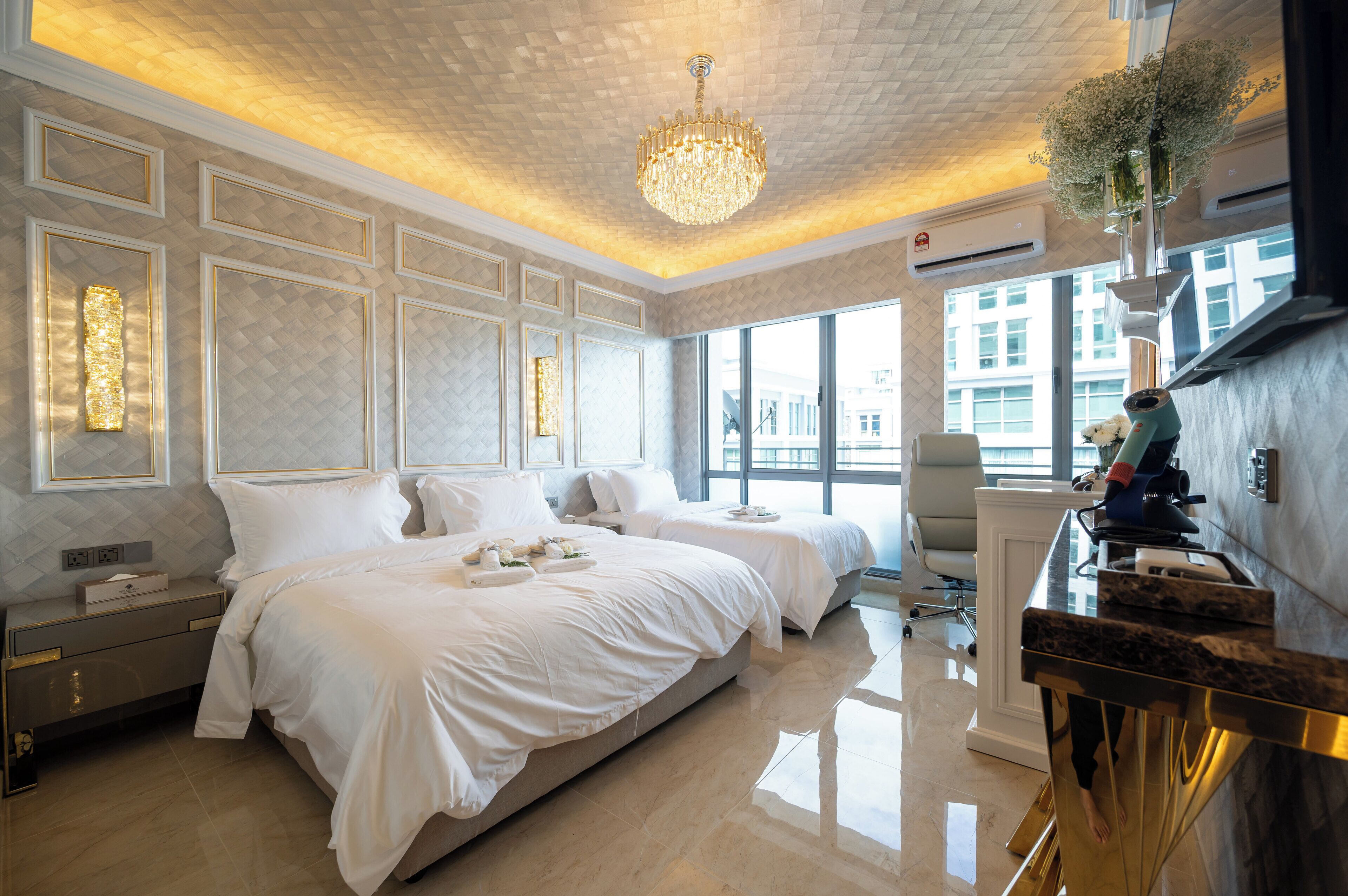 A modern hotel room with two beds, a large window, a chandelier, and a desk with a computer.