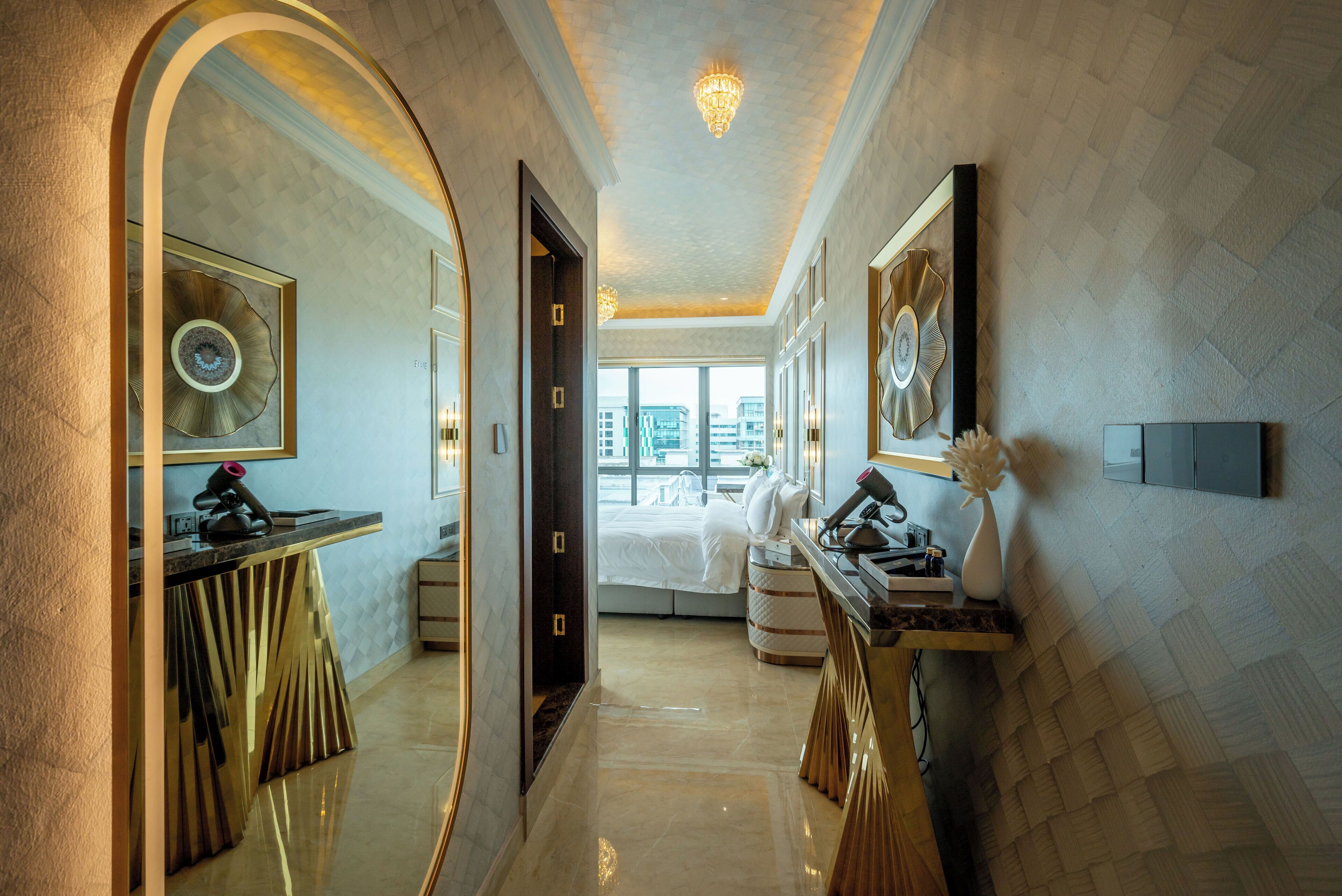 A hallway with a mirror, a vanity, and a bed in the background.