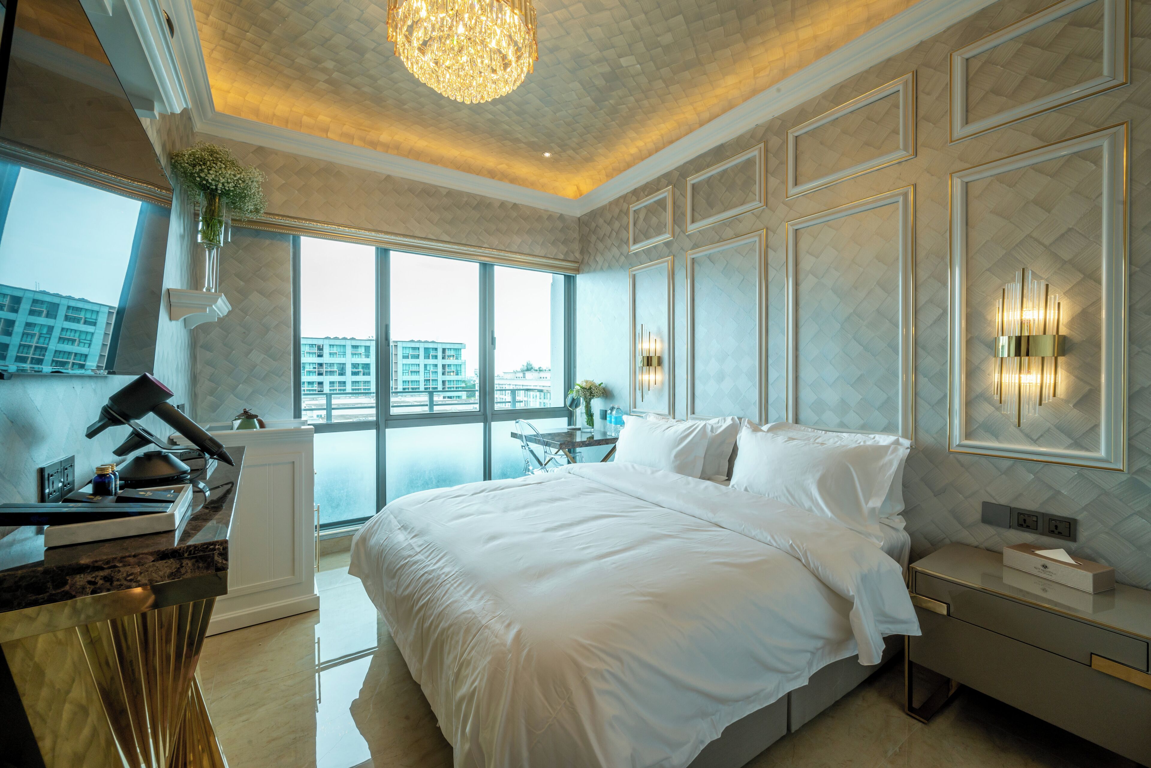 A modern hotel room with a large bed, a desk with a lamp, a view of the city through large windows, and decorative wall sconces.