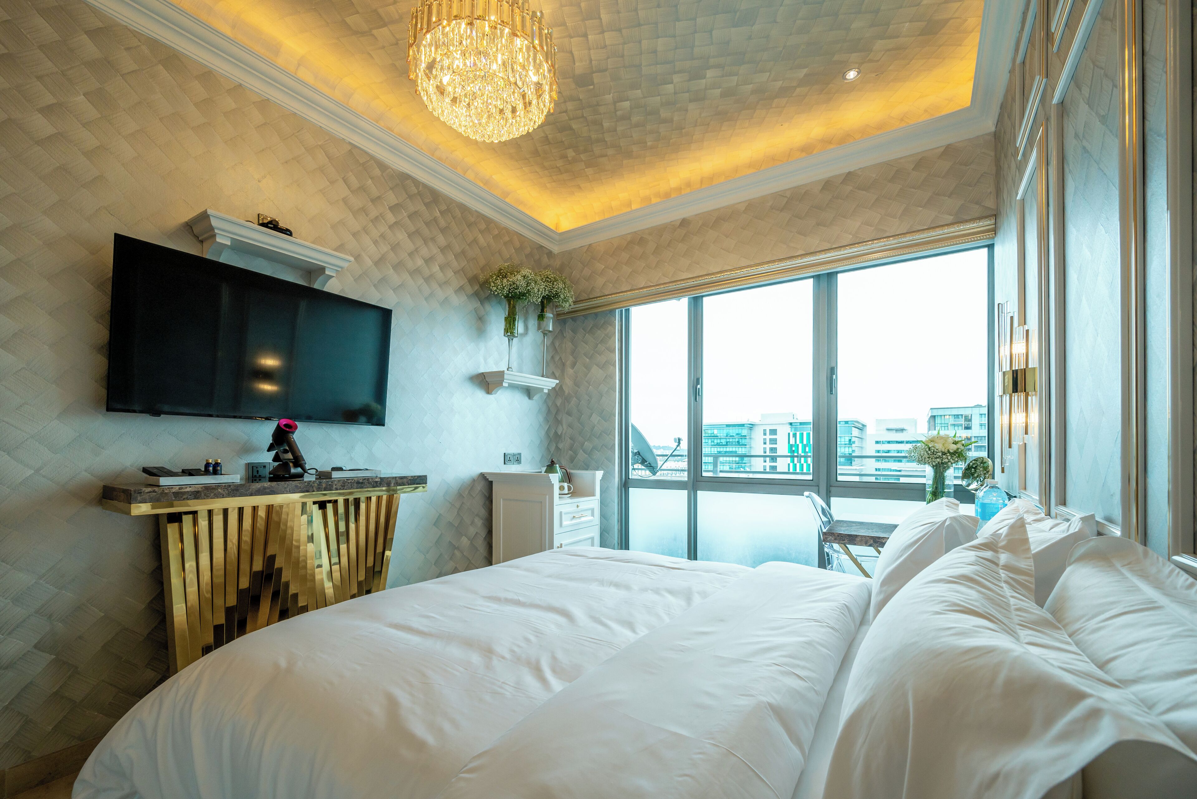 Een moderne hotelkamer met een groot bed, een wandtelevisie, een houten console en uitzicht op de stad door grote ramen.