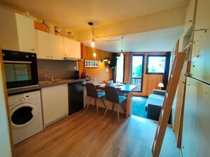 Een kleine keuken met houten kastjes, een inbouwoven, een wasmachine en een eethoek met een tafel en stoelen.