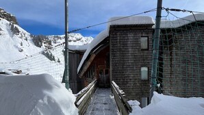 A snow-covered pathway leading to a wooden building with a metal fence and snowbanks on either side.