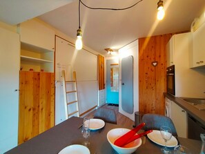 Een praktische keuken met een koelkast, fornuis en houten details. Een eettafel met stoelen en een ladder zijn te zien.