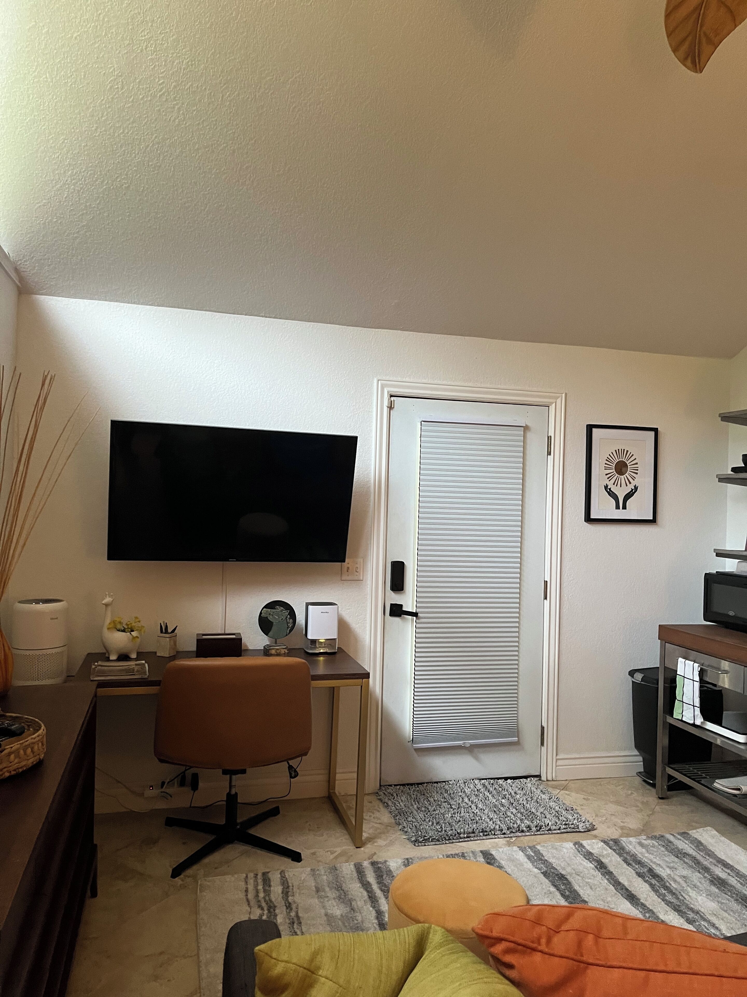 Un dormitorio con cama, escritorio con computadora, televisión, puerta y un cuadro enmarcado en la pared.