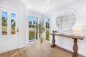 A room with a wooden floor, a round mirror, a wooden console table with a lamp, and a view of a garden through a glass door.