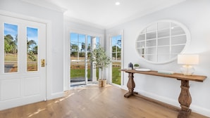 A room with a wooden floor, white walls, a round mirror, a wooden console table with a lamp, and a door leading to a garden.