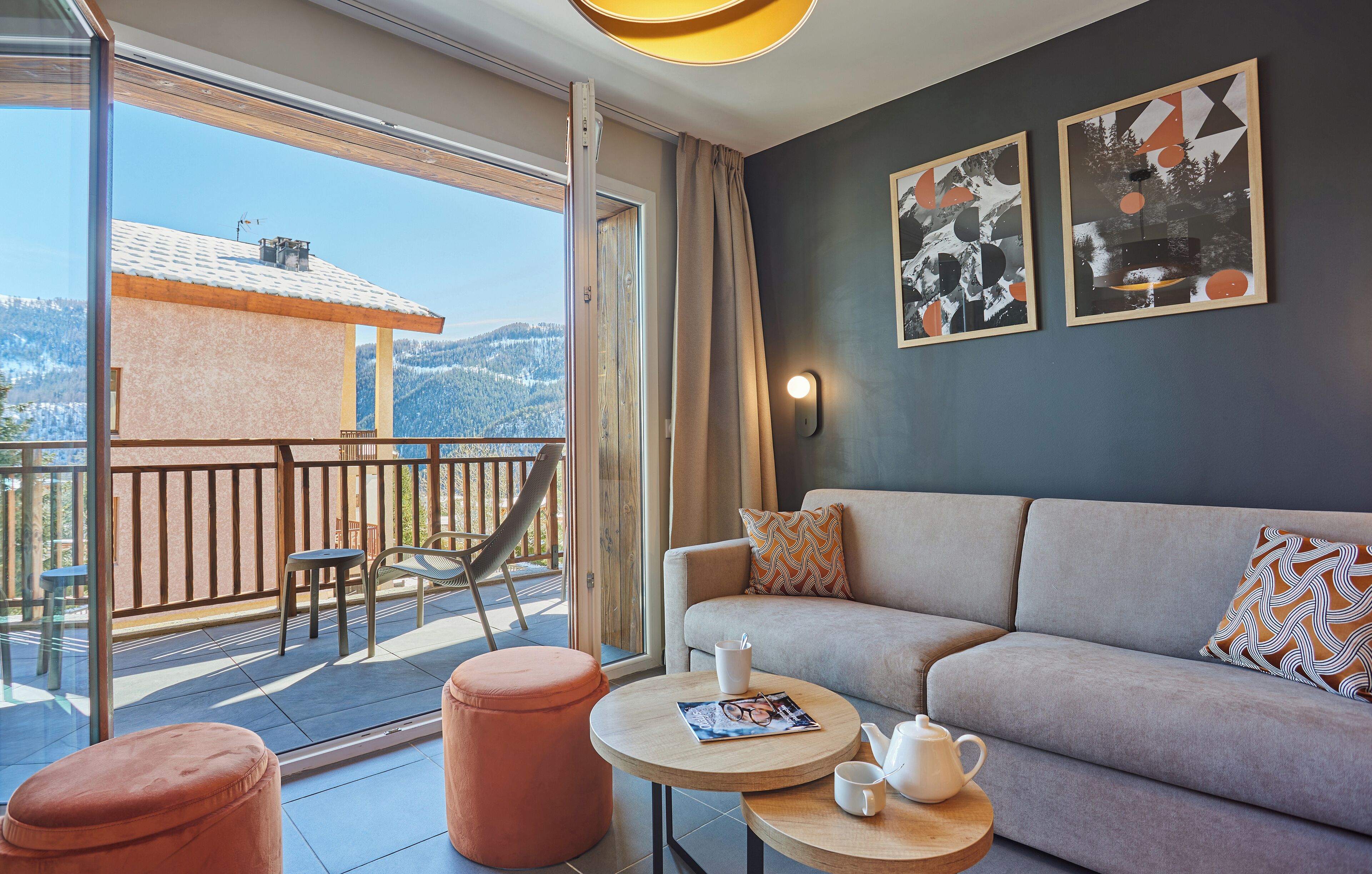 Un salon moderne comprenant un canapé, une table basse et un pouf, un balcon offrant une vue sur les montagnes, ainsi que deux œuvres d'art abstraites accrochées au mur.