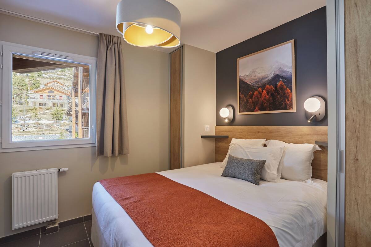 A hotel room with a bed, a window with curtains, a radiator, a wall-mounted light, and a framed picture.