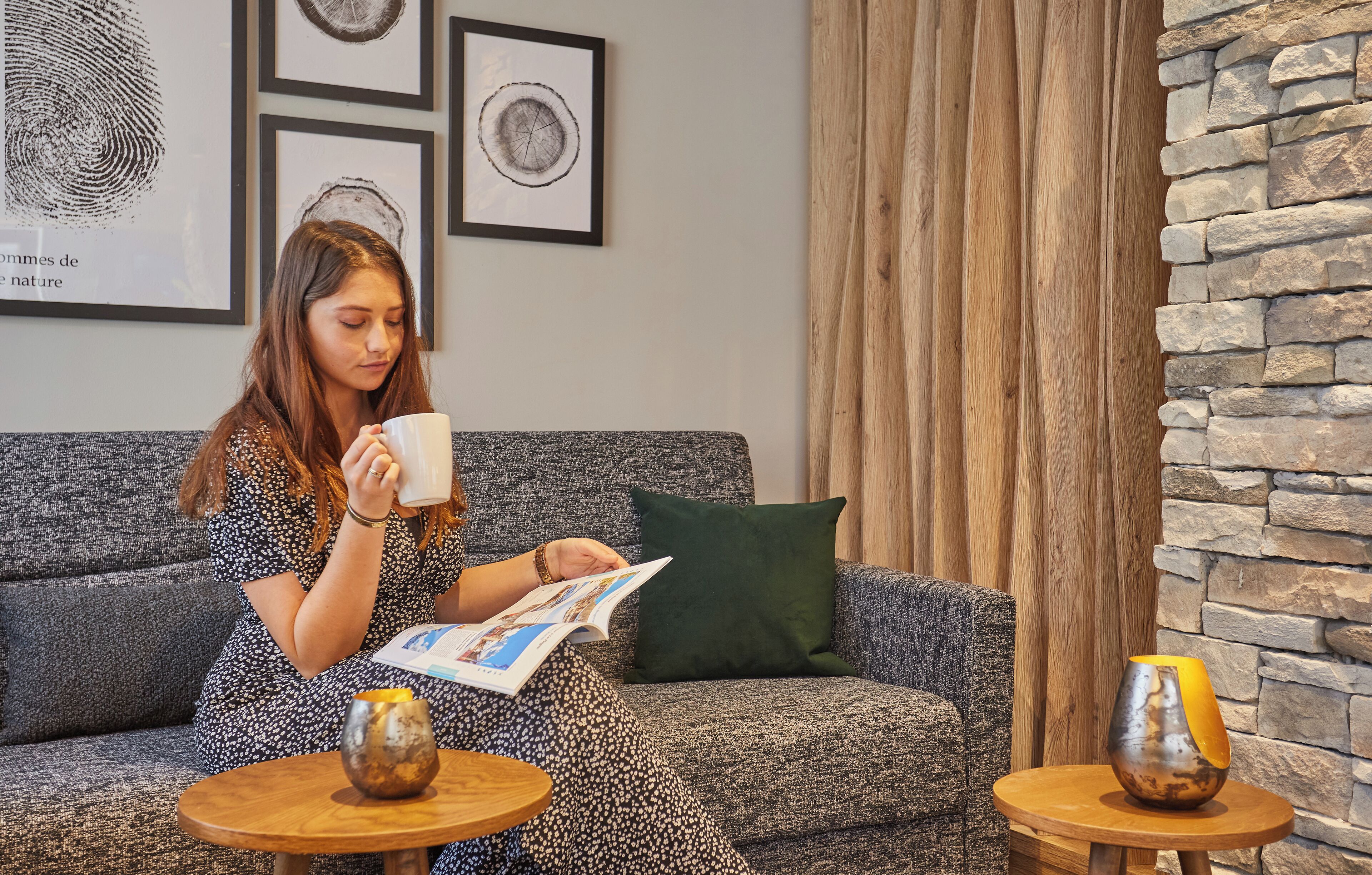 Una persona sentada en un sofá gris, con una revista, una taza blanca y un cojín verde. Hay dos mesitas redondas con jarrones decorativos.
