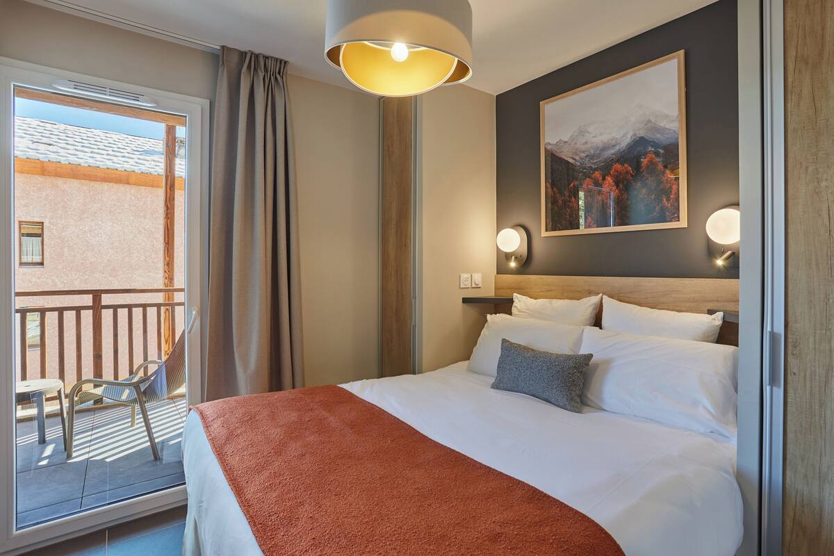 A hotel room with a bed, a large window, a balcony, and a painting on the wall.