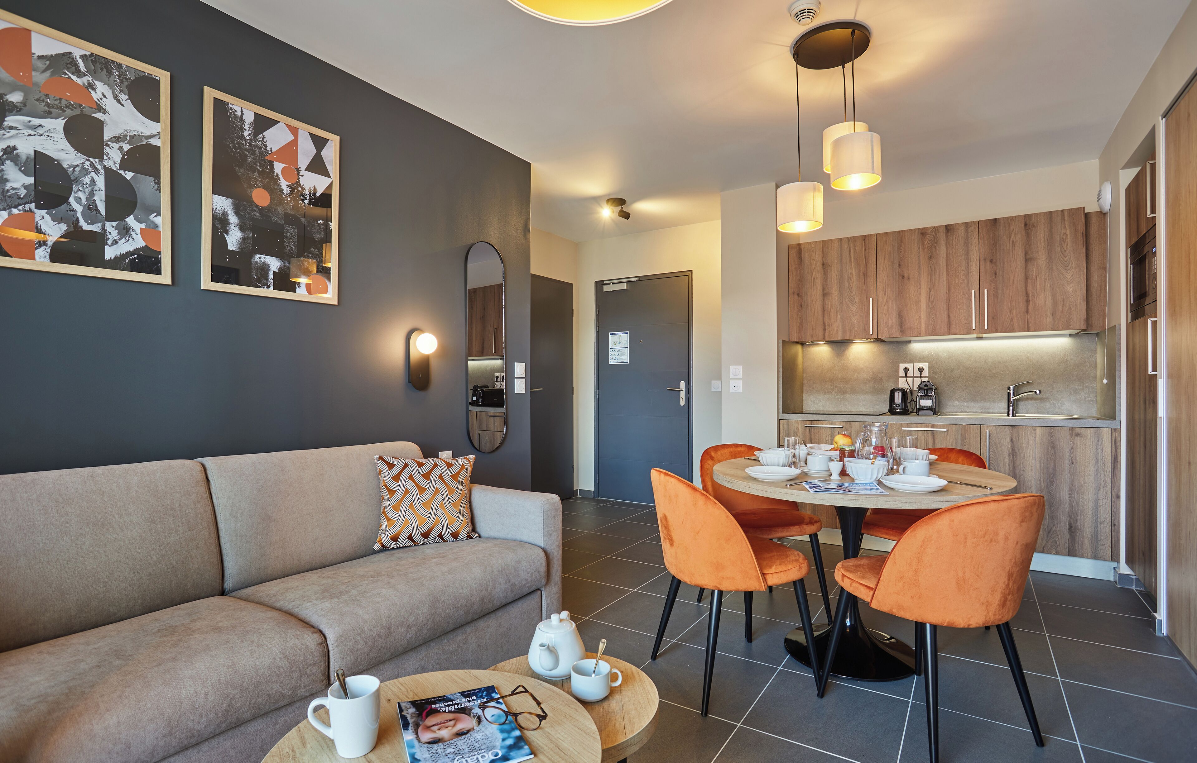 A modern living room with a sofa, coffee table, and dining area with chairs. There is a kitchen in the background with wooden cabinets and a countertop.