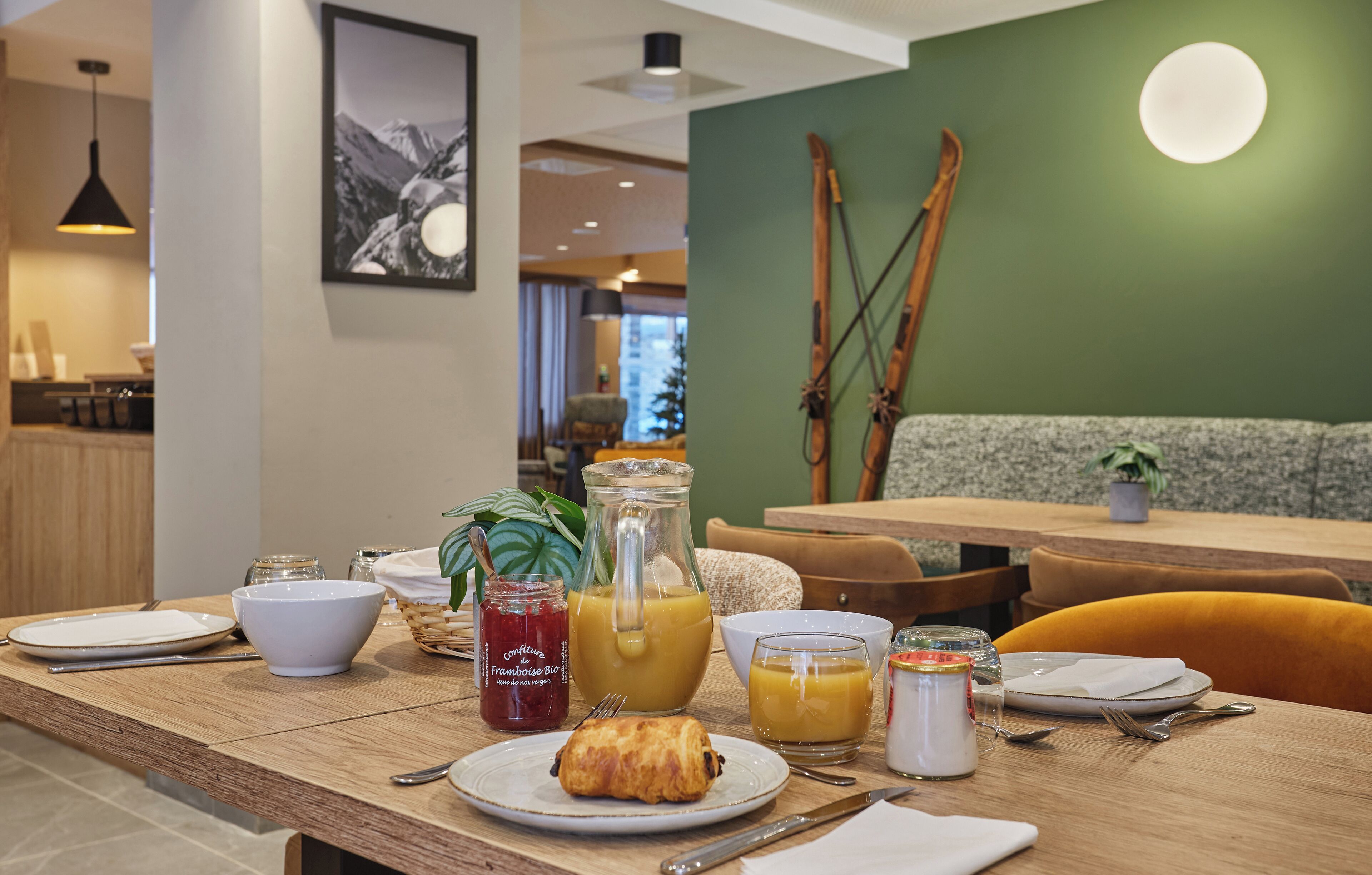 Ein Frühstückstisch mit Holzoberfläche, Tellern, einem Croissant, einer Karaffe Orangensaft und einer Schale Marmelade.