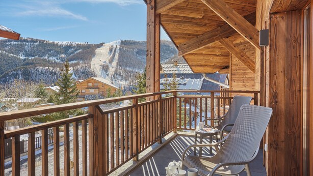Un balcón de madera con dos sillas y una mesa, con vistas a un paisaje montañoso nevado.