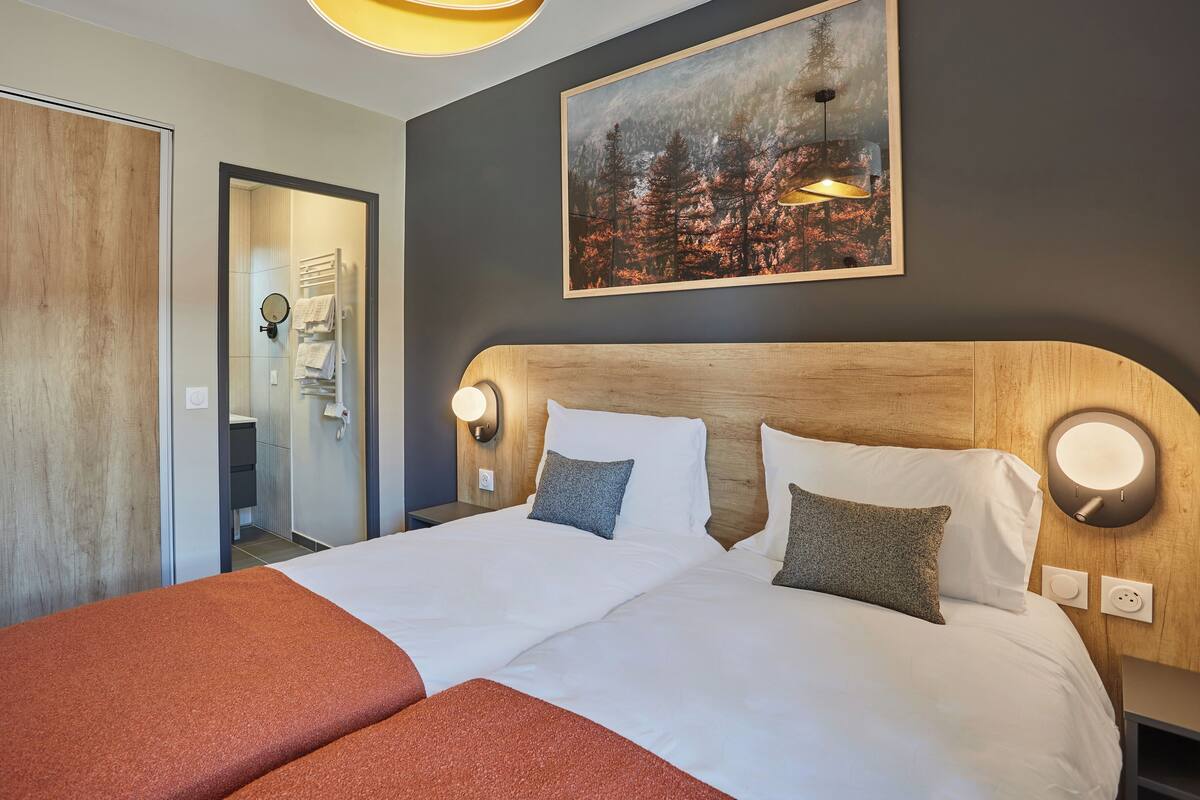 A hotel room with a large bed, a wooden headboard, a painting of a forest, and a wall-mounted light fixture.