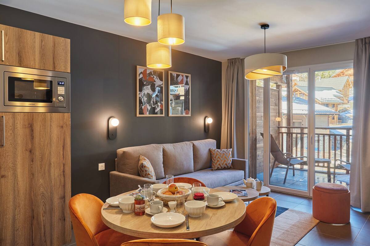 A modern kitchen with a built-in microwave, wooden cabinets, and a dining area with orange chairs and a round table set for breakfast.
