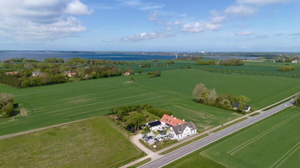 Vista aérea de una zona rural con campos verdes, una casa grande de color blanco y techo rojo, y una carretera que serpentea paralela a los campos.