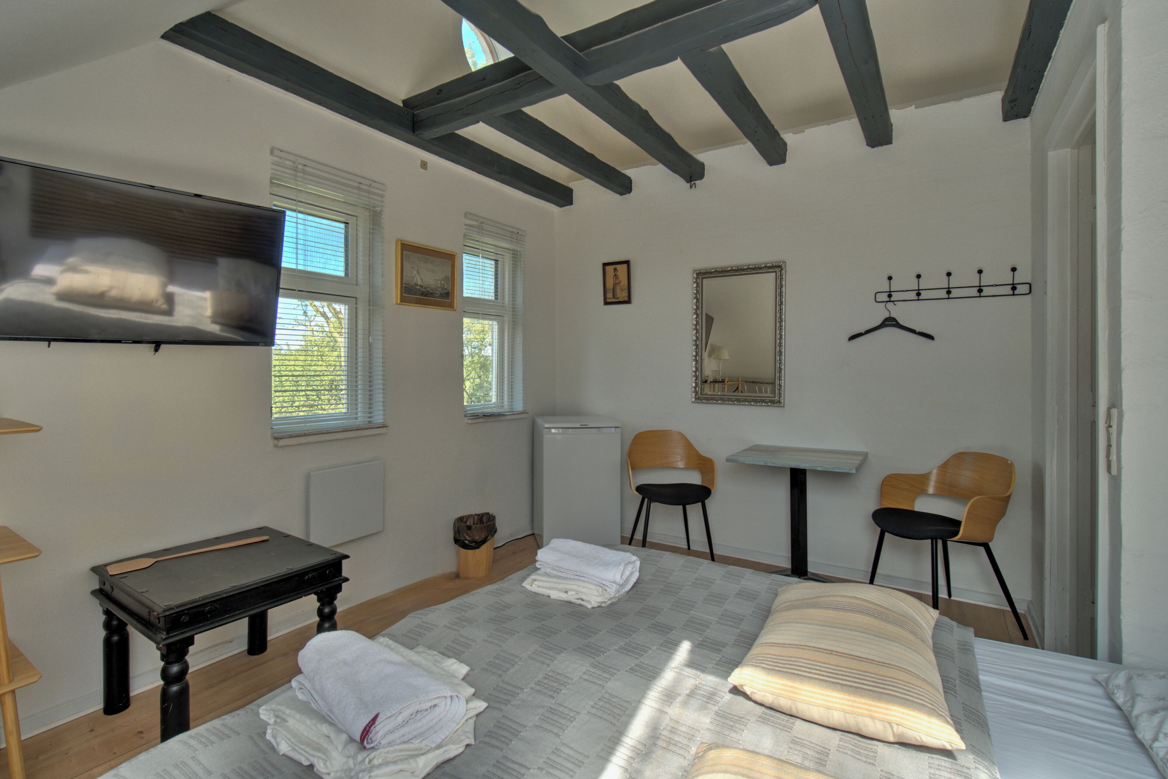 Une chambre à coucher comprenant un lit, une télévision, une petite table, deux chaises, un miroir et un porte-manteau.