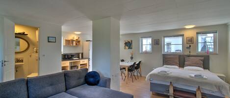A modern apartment with a grey sofa, a bed with a wooden headboard, a dining area, and a kitchen.