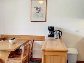 Uma mesa de jantar de madeira com cadeiras, uma tigela branca com abobrinha, uma cafeteira preta e um quadro na parede.