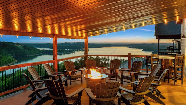 A covered outdoor seating area with Adirondack chairs, a central fire pit, and a view of a lake and mountains.