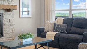 A living room with a stone fireplace, a dark sofa with a 'lake house' pillow, a wooden coffee table with a plant, and a large window with a view of greenery.
