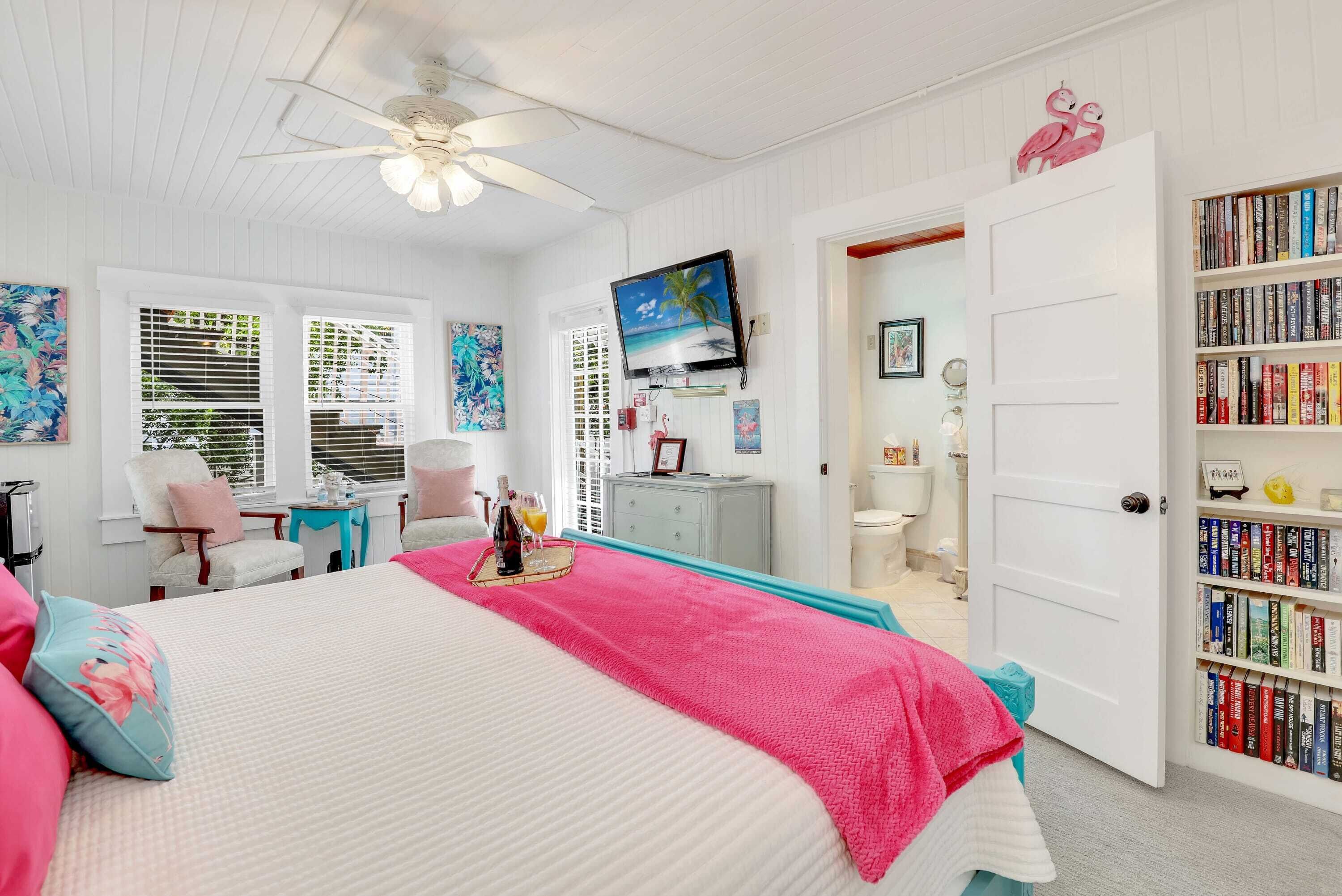 A bedroom with a bed, a ceiling fan, a TV, a bookshelf, and a door leading to another room.