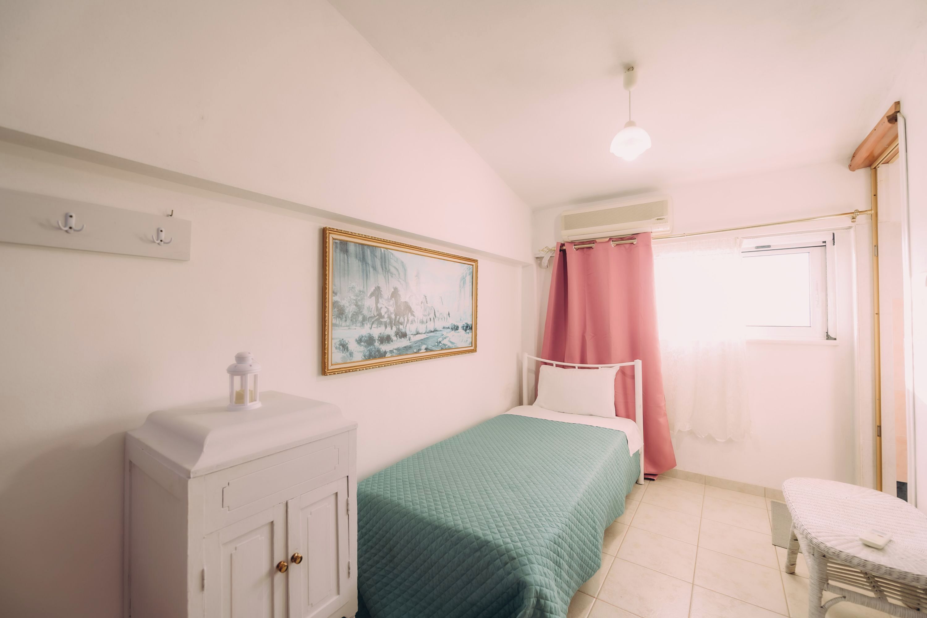 Een slaapkamer met een bed, een witte inbouwkast, een schilderij aan de muur en een raam met roze gordijnen.