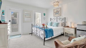 Un dormitorio con estructura de cama metálica, paredes blancas, cómoda de madera, silla de mimbre y mesita de noche con lámpara.